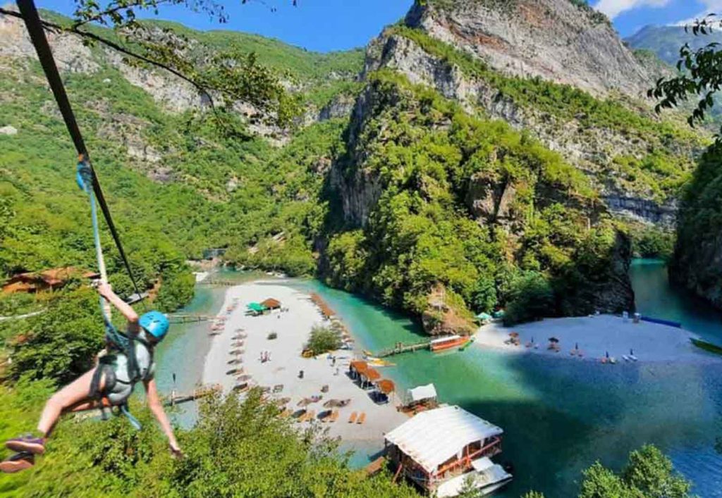 A person ziplining over Shala River, with clear water, green trees, and mountains in the background.